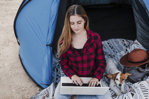 Junge Freiberuflerin mit Laptop neben einem Hund im Zelt sitzend - EBBF04220