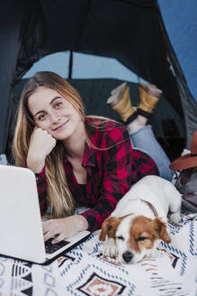 Lächelnde schöne junge digitale Nomadin liegend mit Laptop und Hund im Zelt - EBBF04221