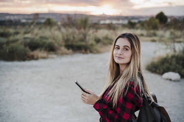 Beautiful young woman wearing backpack holding smart phone during sunset - EBBF04230