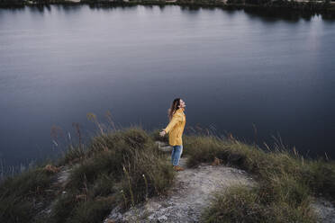 Junge Frau in gelbem Regenmantel steht mit ausgestreckten Armen am Seeufer - EBBF04233