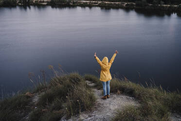 Young woman wearing yellow raincoat standing with arms raised by lake - EBBF04234