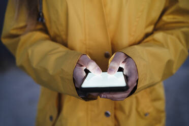 Junge Frau in gelbem Regenmantel, die eine Textnachricht über ihr Smartphone sendet - EBBF04238