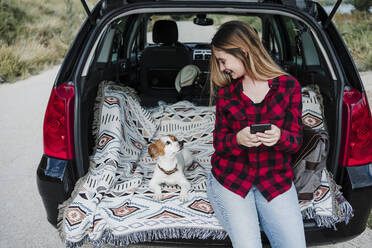 Lächelnde junge Frau mit Smartphone sitzt neben einem Hund im Kofferraum eines Autos - EBBF04239