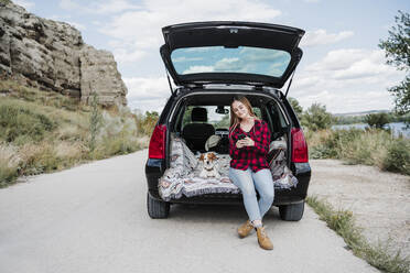 Junge Frau, die ein Mobiltelefon benutzt, während sie neben einem Hund im Kofferraum eines Autos sitzt - EBBF04240