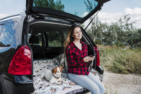 Glückliche Frau, die ein Mobiltelefon hält, sitzt neben einem Hund im Kofferraum eines Autos - EBBF04241