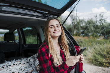 Lächelnde schöne Frau mit blondem Haar, die ein Smartphone im Kofferraum eines Autos hält - EBBF04242