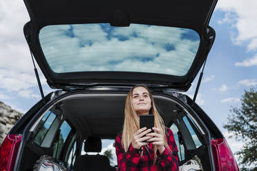 Blonde Frau sitzt mit Smartphone im Kofferraum eines Autos - EBBF04243