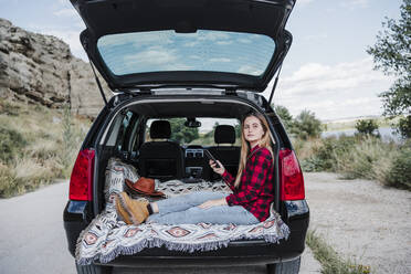 Young blond woman sitting with smart phone in car trunk - EBBF04244