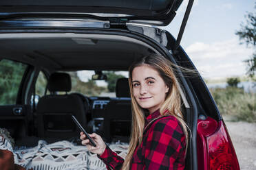 Lächelnde blonde Frau sitzt mit Handy im Kofferraum eines Autos - EBBF04245
