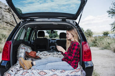 Young woman text messaging through smart phone while sitting in car trunk - EBBF04246