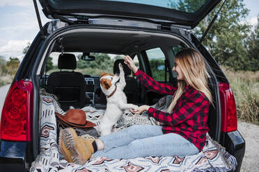 Glückliche junge Frau spielt mit Hund im Kofferraum eines Autos - EBBF04247