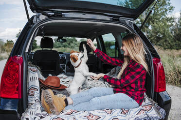 Happy young woman playing with dog in car trunk - EBBF04247