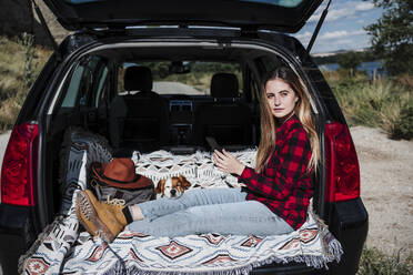 Schöne junge Frau, die wegschaut, während sie mit ihrem Hund im Kofferraum eines Autos an einem sonnigen Tag sitzt - EBBF04248