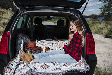 Lächelnde junge Frau, die ein Smartphone benutzt und mit ihrem Hund im Kofferraum eines Autos sitzt, an einem sonnigen Tag - EBBF04249