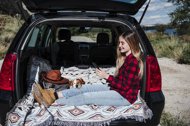 Smiling young woman using smart phone sitting with dog in car trunk on sunny day - EBBF04249