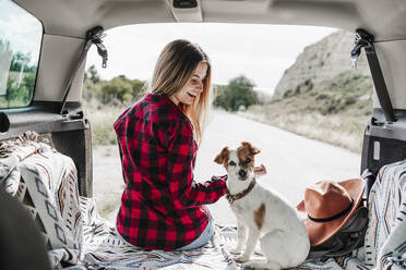 Happy blond woman sitting with dog in car trunk - EBBF04251