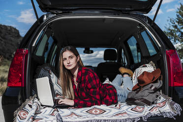 Freiberuflerin liegt mit Laptop im Kofferraum eines Autos an einem sonnigen Tag - EBBF04252