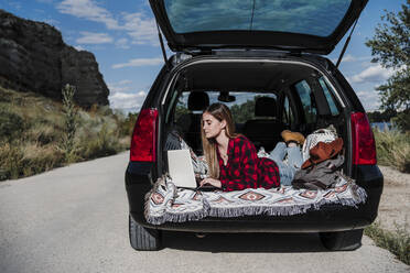 Junge digitale Nomadin mit Laptop im Kofferraum eines Autos an einem sonnigen Tag - EBBF04253