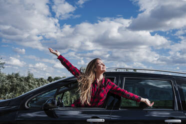 Happy blond woman with arms outstretched leaning from car window during sunny day - EBBF04256