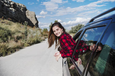 Smiling young woman leaning from car window enjoying sunny day - EBBF04257