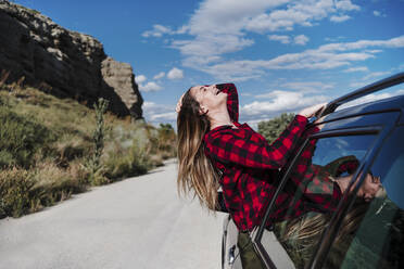 Fröhliche junge Frau mit Hand im Haar, die sich an einem sonnigen Tag aus dem Autofenster lehnt - EBBF04258
