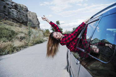 Smiling young woman with hand raised leaning from car window - EBBF04259