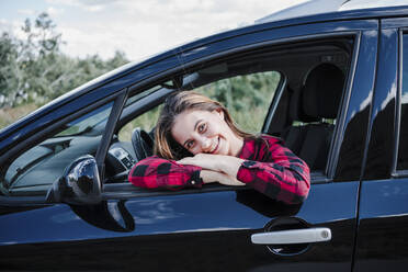 Glückliche junge Frau, die sich im Auto sitzend ans Fenster lehnt - EBBF04260