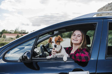 Happy young woman with dog sitting in car - EBBF04263