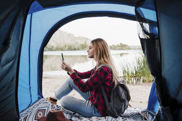 Junge blonde Frau, die im Zelt sitzend eine SMS über ihr Smartphone verschickt - EBBF04268