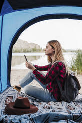 Schöne junge blonde Frau surft im Internet über ihr Smartphone im Zelt - EBBF04269