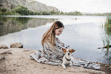 Smiling young woman wrapped in blanket sitting by dog at lakeshore - EBBF04272