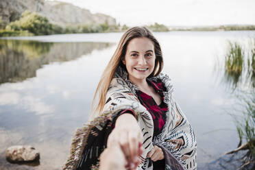 Glückliche junge Frau in eine Decke gewickelt, die die Hand einer Freundin am Seeufer hält - EBBF04274