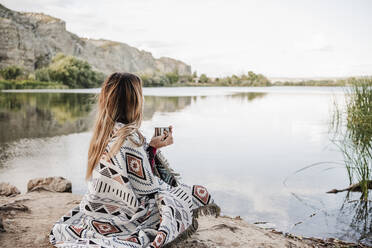 Junge blonde Frau in Decke eingewickelt mit Kaffeetasse am Seeufer - EBBF04276