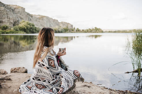 Young blond woman wrapped in blanket holding coffee mug at lakeshore - EBBF04276