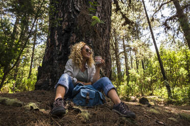 Smiling woman holding coffee mug while sitting in forest - SIPF02310