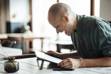 Glücklicher glatzköpfiger Geschäftsmann, der in einem Café sitzt und auf ein digitales Tablet schaut - GUSF06081
