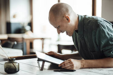 Happy bald businessman looking at digital tablet while sitting in cafe - GUSF06081