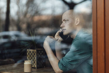 Businessman looking through window while talking on mobile phone at cafe - GUSF06114