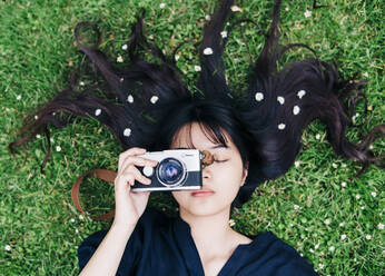 Weibliche Fotografin mit Blumen im Haar hält die Kamera, während sie im Gras liegt - ASGF00794
