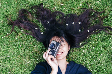 Glückliche Fotografin mit Blumen im Haar, die die Kamera hält, während sie im Gras liegt - ASGF00795