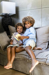 Afro brothers with book sitting on sofa at home - DLTSF01993