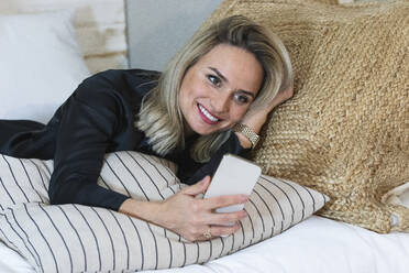 Nachdenkliche Geschäftsfrau, die ihr Smartphone hält, während sie zu Hause auf dem Bett liegt - PNAF02028