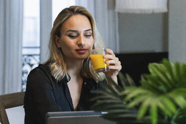 Businesswoman holding juice glass while looking at digital tablet at home - PNAF02037