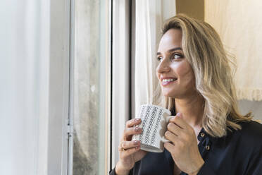 Nachdenkliche Geschäftsfrau, die eine Tasse in der Hand hält, während sie durch das Fenster ihres Hauses schaut - PNAF02042