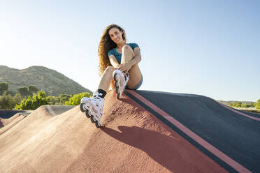 Junge Frau mit braunen Haaren, die Inline-Skates trägt und auf einem Pumptrack sitzt - DLTSF02035