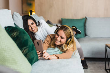 Ein paar lesbische Frauen liegen auf dem Sofa und machen Selbstaufnahmen mit dem Smartphone, während sie am Wochenende im Wohnzimmer zusammen chillen - ADSF25697
