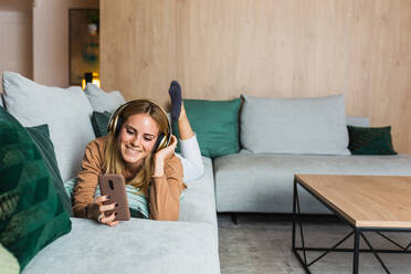 Fröhliche Frau liegt auf dem Sofa und genießt Musik über Kopfhörer, während sie auf den Bildschirm ihres Smartphones schaut - ADSF25698