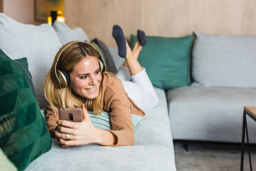 Fröhliche Frau auf dem Sofa liegend und mit Kopfhörern Musik genießend, während sie mit ihrem Smartphone wegschaut - ADSF25699