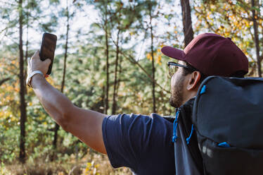 Side view of young ethnic male hiker in casual wear and cap with backpack taking a self portrait with his mobile phone in lush summer woodland - ADSF25731