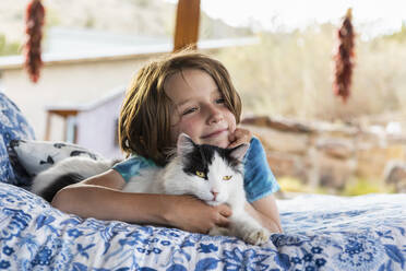 Young boy lying on outdoor bed stroking a pet cat - MINF16278